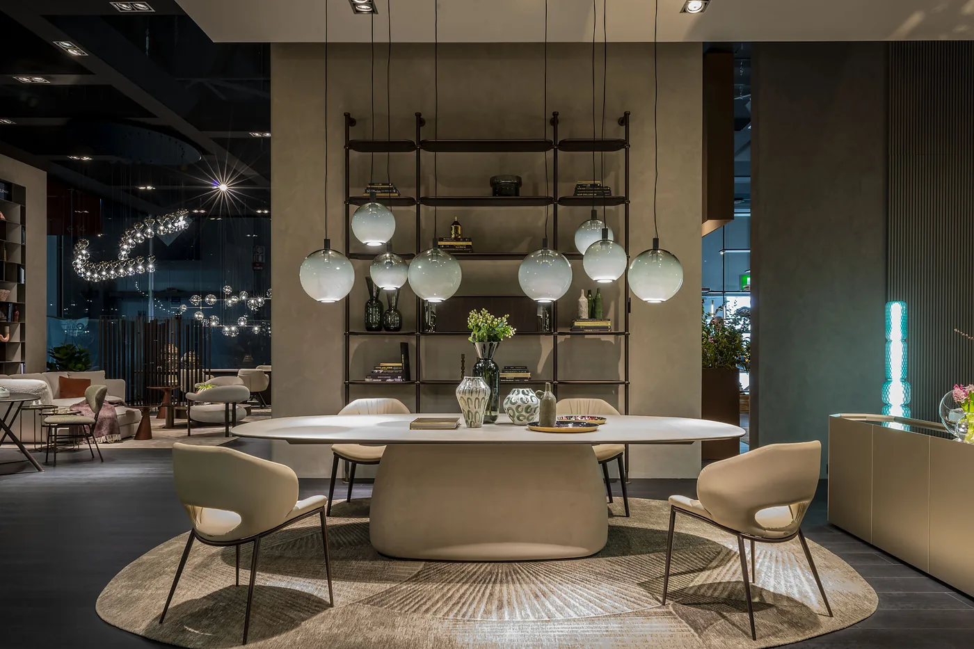 Grande table ovale en céramique claire sur pied central, chaises grises, dans une salle sombre ouverte sur une baie vitrée