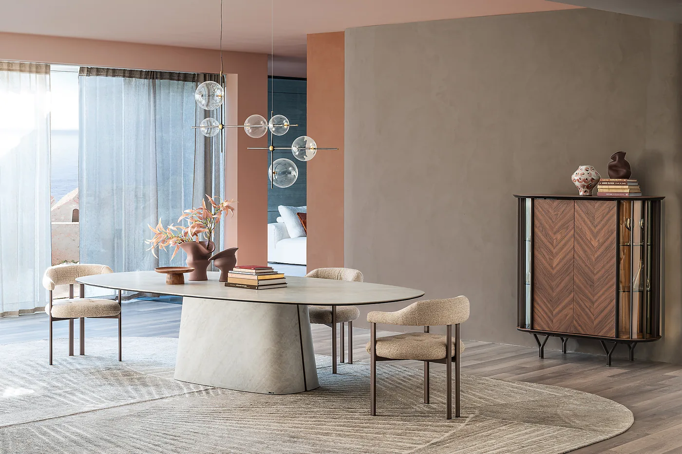 Table ovale en céramique claire sur pied central, fauteuils écrus, devant de grandes fenêtres