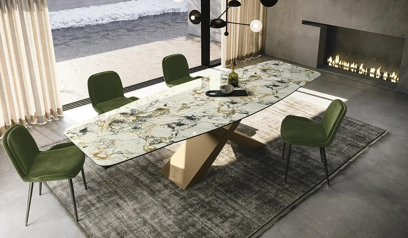 Table au plateau céramique marbré sur piètement croisé doré, chaises en velours vert, près d’une cheminée