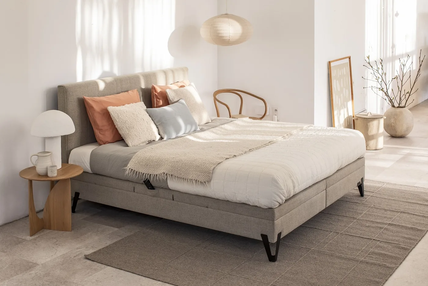 Boxspring en tissu beige aux pieds noirs, coussins orangés et plaid crème, lampe sur un chevet en bois