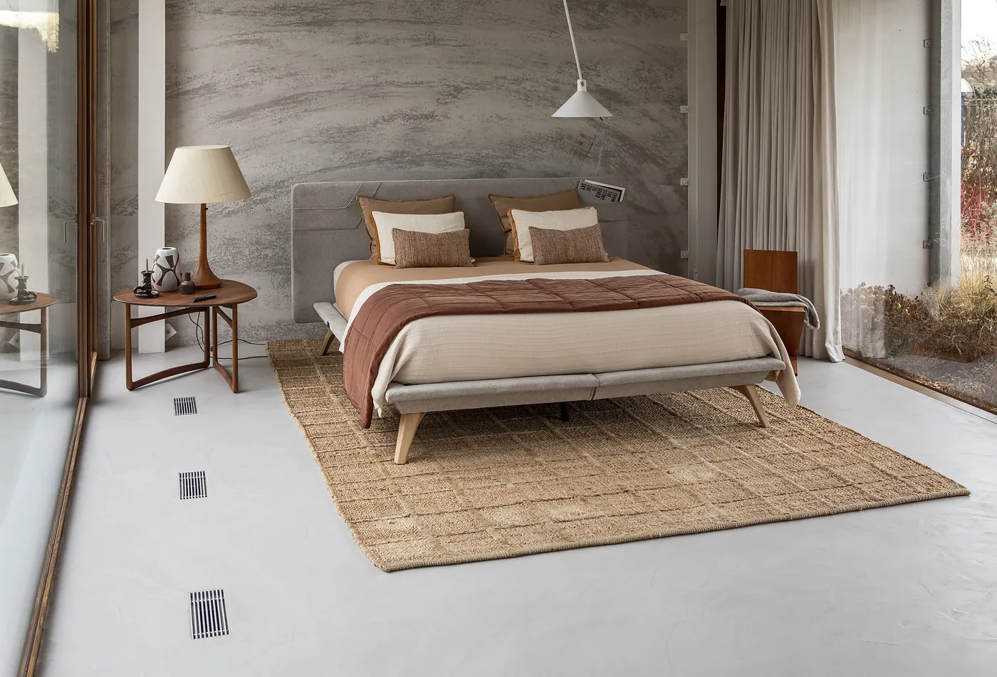 Chambre au mur de pierre claire : lit tapissé gris sur pieds en bois, linge ocre et brique, tapis tressé